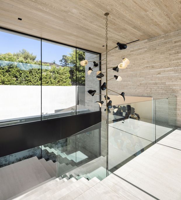Modern interior landing overlooking a stairwell, with pale timber-clad ceiling and flooring, textured light stone or brick wall finish, extensive floor-to-ceiling glazing, glass balustrades, and a sculptural black-and-white pendant light. The open, sunlit space overlooks leafy landscaping outside and features a dramatic stair void.