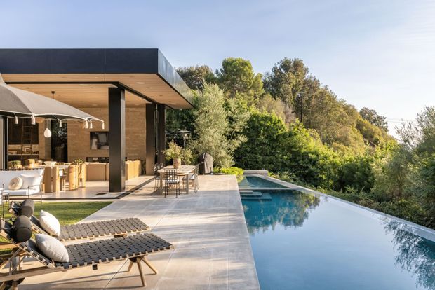 A contemporary flat-roofed house opens onto a broad stone-paved terrace with an infinity-edge swimming pool. The open-plan kitchen and living area is visible beneath a warm timber-lined soffit, supported by dark slender columns and framed by extensive glazing. Outdoor loungers and a dining setting occupy the terrace, with dense landscaped greenery beyond.