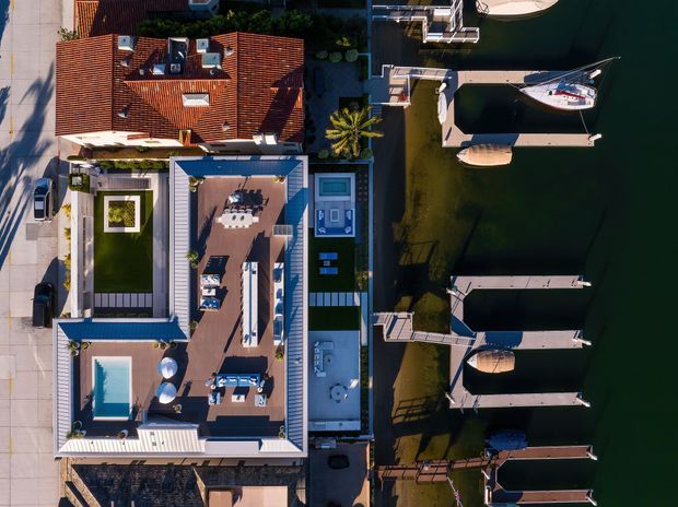 Aerial top-down view of a waterfront residential property with several connected or adjacent flat-roofed structures, terracotta-tiled roofing, landscaped courtyards, rooftop terraces, a swimming pool, and private boat docks extending into calm dark water. The image emphasizes the relationship between the home, outdoor living spaces, pool, and marina-like waterfront setting.