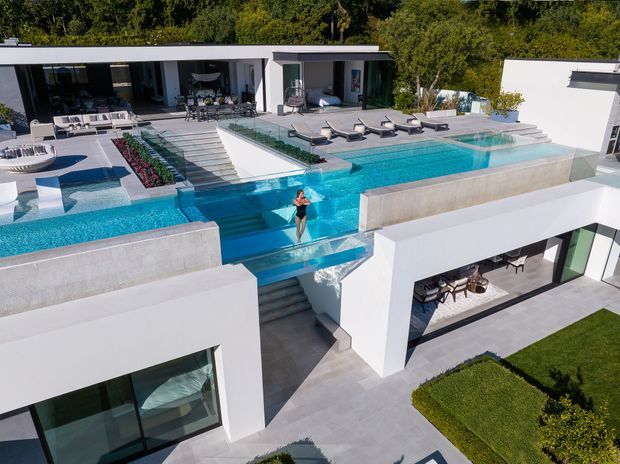 Aerial view of a large contemporary white residence arranged around multiple elevated terraces and a vivid turquoise swimming pool. The property features flat rooflines, expansive dark-framed glazing, broad concrete or stone-paved terraces, outdoor dining and lounge areas, sun loungers, planted garden beds, and a dramatic raised pool with spillover water and stairs connecting the levels. Dense trees form the background.