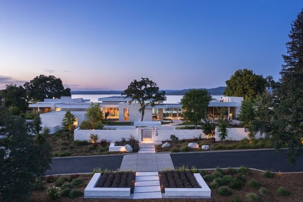 Elevated dusk view of a low, sprawling modern residence with crisp white rendered volumes, extensive horizontal glazing, flat rooflines, terraces, and a formal landscaped entry. The property sits beside a broad body of water with distant hills visible beyond, and is approached by a paved path across a landscaped front garden.
