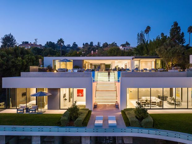 Dusk exterior view of a large, symmetrical contemporary residence set into a landscaped suburban hillside. The white and pale grey flat-roofed facade features extensive floor-to-ceiling glazing, illuminated open-plan living areas, broad terraces, a dramatic central staircase, manicured lawns, and a curved foreground structure or pool edge. Tall palms and mature vegetation surround the property.
