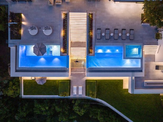 Aerial dusk view of a landscaped residential outdoor area featuring two illuminated blue swimming pools, a central linear water feature or stair-like pool connection, paved entertaining terraces with outdoor dining and lounge furniture, planted garden beds, manicured lawns, and curved boundary landscaping. The composition emphasizes the pool area and overall outdoor design.