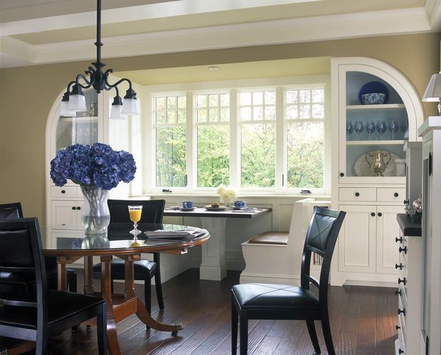 Elegant formal dining room with a round dark wood dining table, black leather dining chairs, built-in white display cabinetry, a bay of multi-pane windows, and a traditional decorative chandelier. The room features dark timber flooring, pale neutral walls, classic moulded trim, and a bright garden outlook.