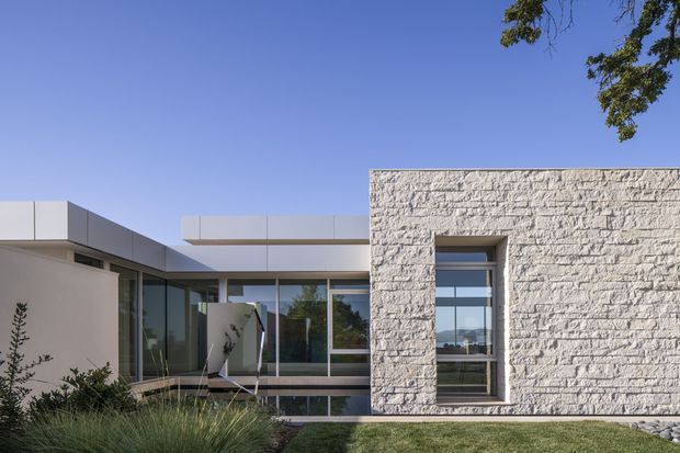Exterior view of a contemporary single-storey residence with a low, flat roofline, extensive floor-to-ceiling glazing, and a prominent pale textured masonry feature wall containing a narrow recessed window. The building is set in landscaped grounds under a clear blue sky, with interior spaces partially visible through the glass.