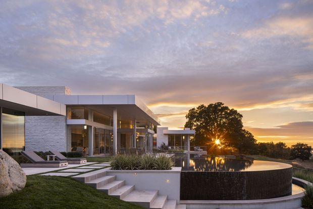 Contemporary multi-level residence viewed from the landscaped grounds at sunset. The building features crisp horizontal roof planes, extensive floor-to-ceiling glazing, pale stone or rendered wall surfaces, and a terrace overlooking a dark-edged reflecting or swimming pool. Concrete steps, clipped lawn, ornamental grasses, and a mature tree frame the scene.