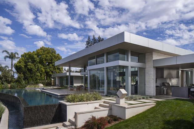 Exterior view of a large contemporary single-storey residence with broad flat roof planes, extensive floor-to-ceiling glazing, pale stone-clad walls, and a sheltered outdoor living area. Landscaped terraces and steps lead to a dark-edged swimming pool, with established trees and a bright partly cloudy sky surrounding the property.