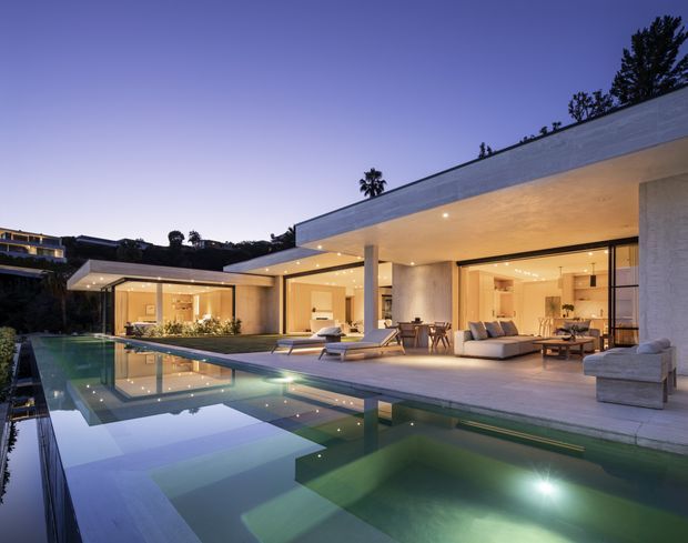 Twilight view of a luxurious, low-slung modern residence organized around a large reflecting swimming pool. The open-plan living area has extensive floor-to-ceiling glazing, broad roof overhangs, recessed lighting, pale stone or rendered walls, and a generous paved terrace with outdoor loungers and seating. Warm interior lighting contrasts with the cool blue evening sky and water reflections.