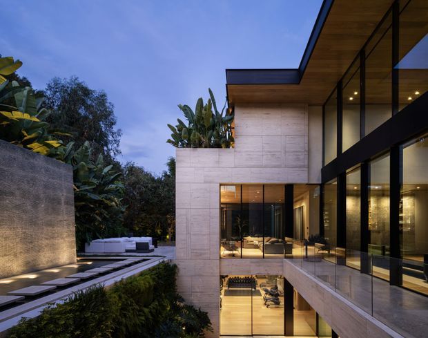 Twilight exterior view of a contemporary multi-level residence set into a lush landscaped site. The home features pale stone or concrete wall planes, extensive black-framed floor-to-ceiling glazing, projecting flat roof planes with warm timber soffits, and planted terraces. Interior living spaces are visible through the glazing, while a stepped path, outdoor seating area, and dense tropical planting occupy the foreground.