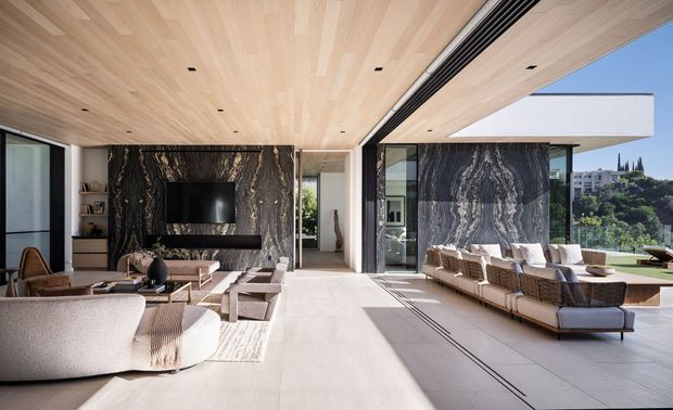 A spacious, open-plan contemporary living room with floor-to-ceiling glazing opening toward an outdoor terrace. The room features a dramatic dark veined stone TV wall and matching feature wall, pale large-format flooring, light timber slatted ceiling, minimalist recessed lighting, and coordinated neutral sofas and lounge chairs. The space is bright, refined, and strongly connected to the outdoors.