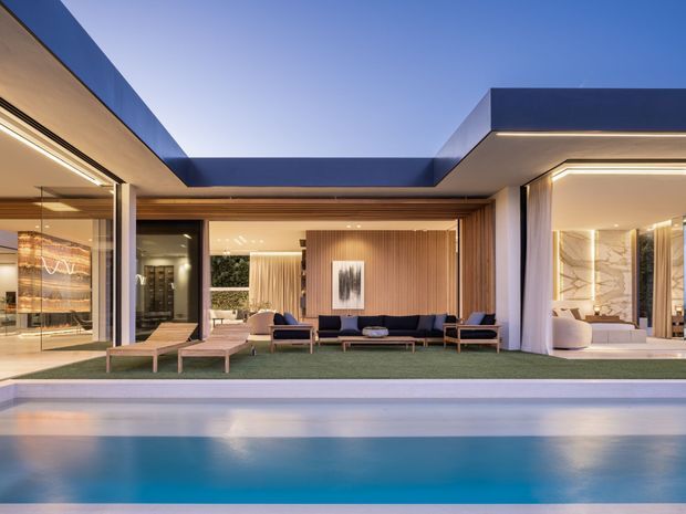 Contemporary luxury residence with a low, flat-roofed composition opening onto a swimming pool. The image looks from the pool toward a covered outdoor living area and glazed interior spaces, with strong indoor-outdoor connection, warm concealed lighting, timber-look vertical wall panelling, pale stone surfaces, and dark roof and frame elements.