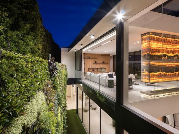 Night-time view of a luxurious contemporary multi-level residence, seen from a landscaped exterior walkway or terrace. Floor-to-ceiling glazing reveals an open-plan living area with pale upholstered seating, warm timber built-in shelving, and a dramatic illuminated amber stone feature pendant or wall element. The home has strong black structural framing, crisp white surfaces, integrated recessed lighting, and lush vertical planting along the edge.