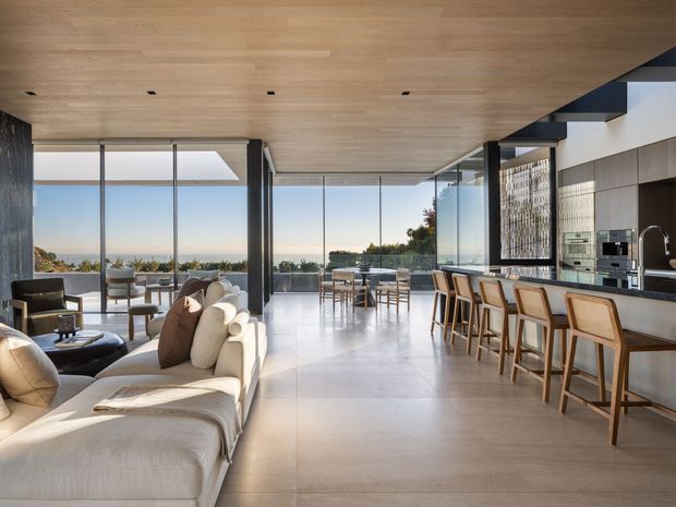 A spacious, light-filled open-plan living, dining and kitchen interior with expansive floor-to-ceiling glazing overlooking a coastal landscape. The room features a pale timber-lined ceiling, large-format light flooring, a sculptural cream sectional sofa, dining furniture, and a long kitchen island with timber-framed bar stools. Dark structural elements and cabinetry create contrast against the warm neutral palette.