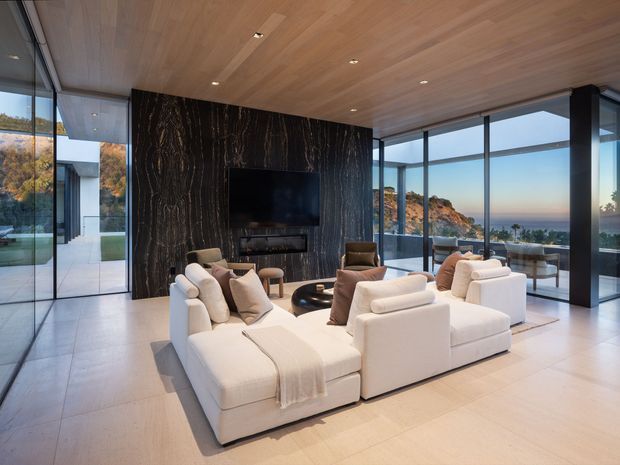 A luxurious contemporary living room with large cream sectional seating arranged around a low dark coffee table and wall-mounted television. The room features expansive floor-to-ceiling glazing, a dramatic dark veined feature wall, pale large-format flooring, and a warm timber-lined ceiling. An adjoining glazed dining or outdoor seating area overlooks a coastal landscape and ocean.