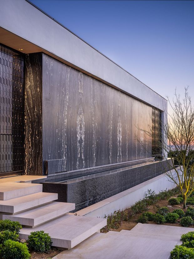 Exterior view of a sleek contemporary flat-roof residence at dusk, featuring a long linear facade clad in dark vertically veined stone or metal panels, pale smooth wall surfaces, a tall patterned metal entry screen, broad floating stone steps, and a landscaped approach with clipped shrubs and bare ornamental trees.