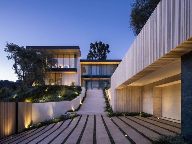 Twilight view of a contemporary multi-level residence on a landscaped sloping site. A broad paved driveway with narrow planted joints leads toward a raised entry stair, flanked by a sculptural white rendered retaining wall and a long timber-clad garage or lower-level structure. The house features extensive floor-to-ceiling glazing, strong horizontal roof planes, vertical timber screening, integrated linear lighting, and mature planting.