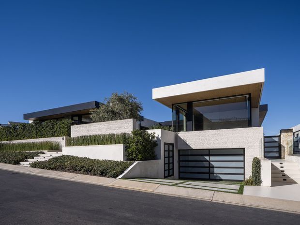 Street-facing view of a contemporary multi-level residence with crisp geometric volumes, a projecting flat roof, extensive dark-framed glazing, pale textured masonry walls, landscaped terraces, and an integrated double garage. The property sits on a sloping suburban site with stepped entry paths and low-maintenance hedging.