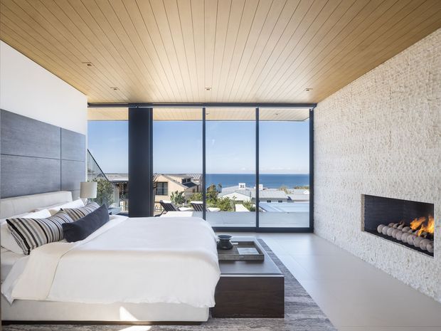 A spacious, refined bedroom with a low-profile white bed, dark wood bedside table, and a contemporary linear fireplace set into a textured white stone feature wall. The room has a warm timber-panelled ceiling, large floor-to-ceiling black-framed glazing, pale flooring, and expansive ocean views beyond the terrace and neighbouring rooftops.