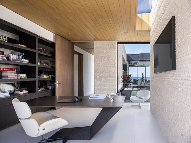 Contemporary home office with a sculptural dark desk, white office chair, extensive built-in black shelving, warm timber-lined ceiling, textured white masonry walls, and polished light flooring. Glazing opens toward a bright courtyard or outdoor area with additional seating and planting.
