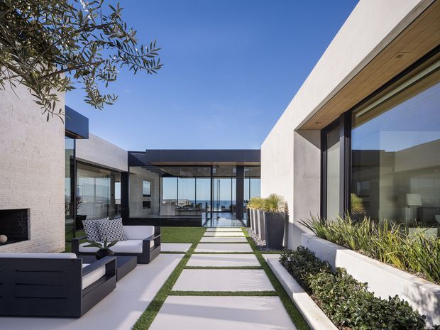 Contemporary low-profile courtyard residence with crisp white rendered walls, extensive floor-to-ceiling glazing, and a linear paved walkway through clipped lawn. The outdoor seating area includes a built-in fireplace wall and dark lounge furniture, while the glazed living space opens toward a distant ocean or horizon view. Clean geometric forms, warm timber soffits, and drought-tolerant planting create a refined indoor-outdoor setting.