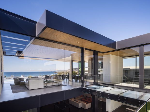A sophisticated contemporary coastal residence with extensive floor-to-ceiling glazing, dark architectural cladding, warm timber soffits, and an open-plan living area overlooking the ocean. The image also reveals a glass-floored bridge or void above a lower level, with sleek outdoor-facing lounge furniture and a bright, expansive setting.