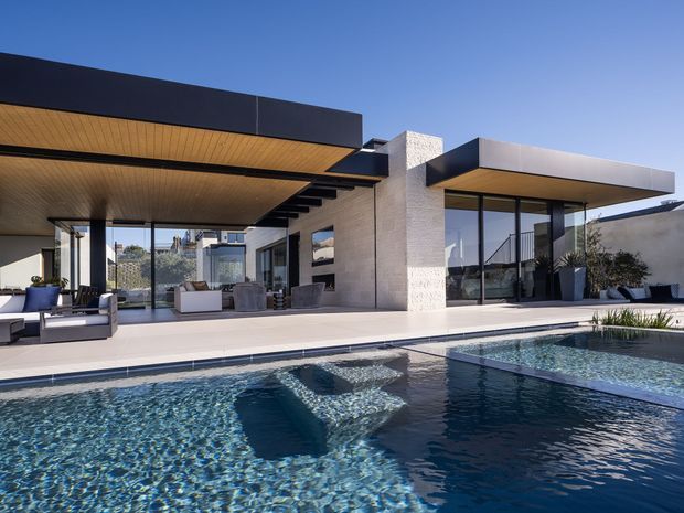 A striking modern single-storey residence arranged around a large swimming pool. The home features broad flat roof planes, extensive floor-to-ceiling glazing, dark metal framing, pale masonry walls, and warm timber soffits. An open-plan living area extends onto a generous pool terrace, with sculptural architectural volumes and a crisp indoor-outdoor connection.