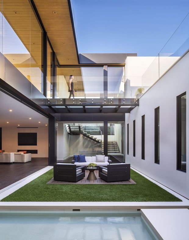 A bright contemporary courtyard viewed from an elevated position, with a shallow reflecting pool in the foreground, artificial lawn and an outdoor lounge seating area at the centre. White rendered walls, extensive floor-to-ceiling glazing, dark metal framing, a glass balustrade and a timber-lined soffit surround the multi-level courtyard. An internal stair and upper-level bridge create a strong open, vertical composition, while an adjacent indoor living area is visible to the left.