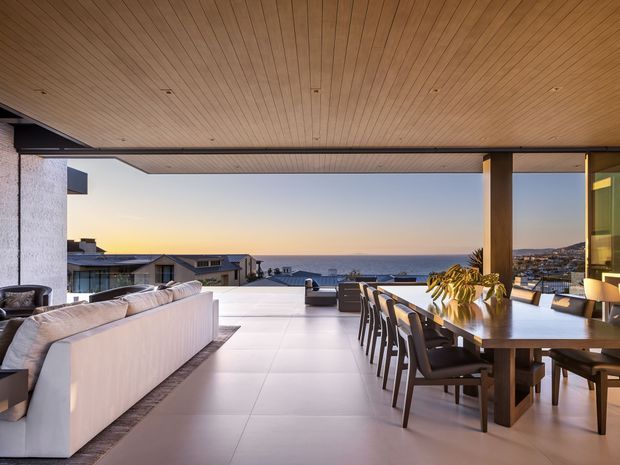 A spacious, open-plan indoor-outdoor living and dining area with a broad ocean-facing view. The room has a warm timber-lined ceiling with recessed downlights, pale large-format floor tiles, a substantial dining table with upholstered chairs, and a large light-colored sectional sofa. Wide openings and slender dark-framed glazing create a seamless connection to the terrace and coastal outlook. The palette is warm and neutral with contemporary, refined styling.