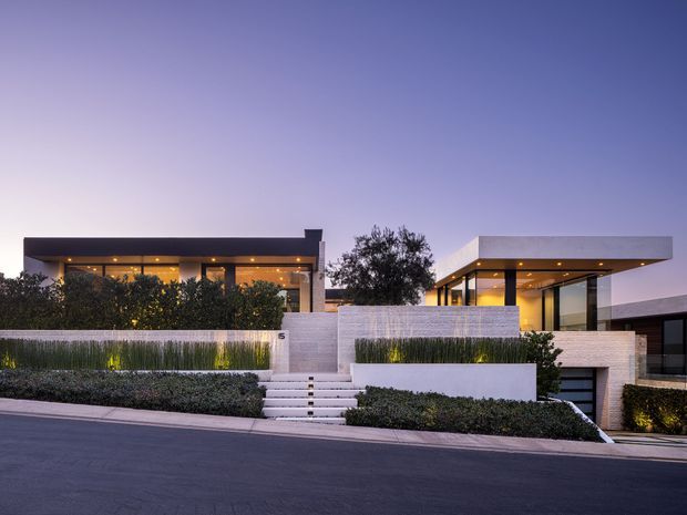 Contemporary flat-roofed residence viewed from across the street at dusk. The home features crisp white masonry walls, extensive glazed openings, deep roof overhangs, recessed exterior lighting, landscaped planting beds, and a stepped entry approach. A darker upper-level pavilion and a partially visible garage are integrated into the modern facade.
