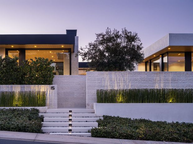Twilight exterior view of a contemporary low-rise residence behind a landscaped front boundary. The home features crisp rectilinear volumes, dark roof planes, extensive floor-to-ceiling glazing, warm recessed lighting, pale masonry walls, and a stepped entry path with integrated lights. Dense shrubs, ornamental grasses, and mature trees soften the modern facade.
