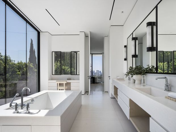 Bright, spacious contemporary bathroom with a freestanding-style built-in white bathtub beside floor-to-ceiling glazing, a long double vanity with integrated basins, large mirrors, black-framed windows, and cylindrical wall lights. The room has pale tiled flooring, white walls and cabinetry, clean linear detailing, and views toward an urban skyline and landscaped surroundings.