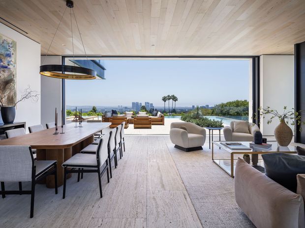 A refined open-plan living and dining space with expansive floor-to-ceiling glazing opening onto a terrace and a panoramic urban landscape. The room features a pale timber ceiling, light natural flooring, a long timber dining table with upholstered chairs, sculptural lounge seating, a low coffee table, minimalist artwork, and restrained contemporary furnishings. The wide opening, strong indoor-outdoor connection, and distant city skyline create an airy, luxurious atmosphere.