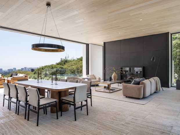Open-plan contemporary living and dining area with floor-to-ceiling glazing overlooking a leafy urban landscape. A large timber dining table with upholstered chairs sits beneath a suspended circular pendant, while a neutral lounge arrangement faces a dark feature wall with a fireplace or media joinery. The space has a light timber-lined ceiling and matching pale timber flooring, creating a refined, airy interior with strong indoor-outdoor connection.