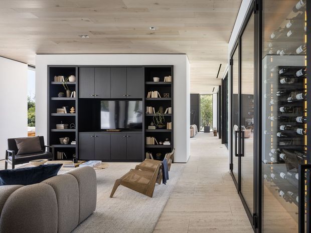 Contemporary open-plan living room with a large black built-in media wall and shelving, neutral upholstered seating, a timber lounge chair, pale timber ceiling, light stone or porcelain flooring, and full-height black-framed glazing. A glass-fronted wine storage cabinet lines the right side of the room, with a bright corridor and outdoor greenery visible beyond.
