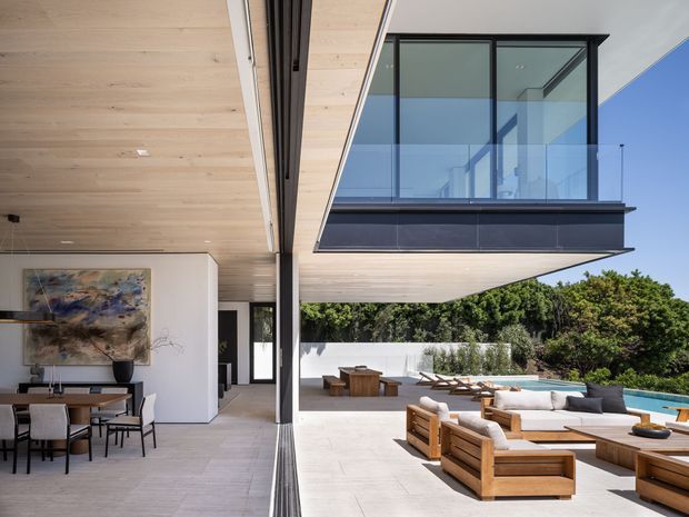 Contemporary indoor-outdoor living and dining area beneath a cantilevered upper level. The space features pale timber ceiling lining, large black-framed glazing, light flooring, a dining setting, timber outdoor lounge furniture, and a swimming pool surrounded by landscaped greenery.