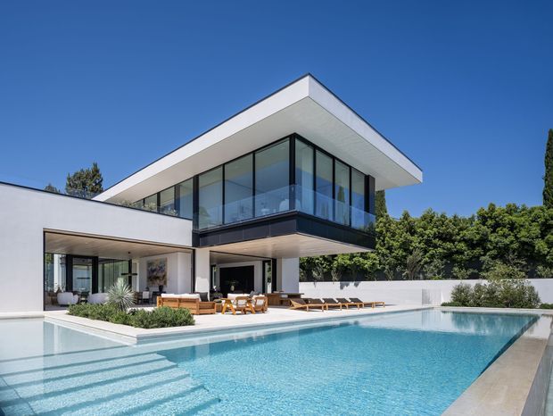 A bright contemporary two-storey residence with crisp white rendered walls, a flat roof and a cantilevered upper-level glass pavilion. The home opens onto a large rectangular swimming pool with a paved edge, outdoor dining and lounge furniture, and landscaped garden screening. Strong indoor-outdoor connections are visible through extensive floor-to-ceiling glazing and covered entertaining areas.