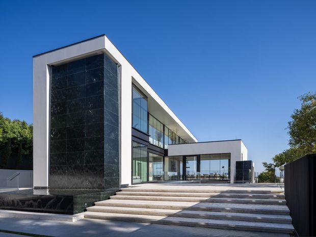 A large contemporary flat-roofed residence viewed from the landscaped forecourt. The crisp white geometric facade incorporates expansive floor-to-ceiling glazing, a tall dark stone feature wall, and broad stepped terraces leading toward an open-plan glass-lined living area. The design is minimalist and strongly horizontal, with a bright blue sky and mature planting surrounding the building.