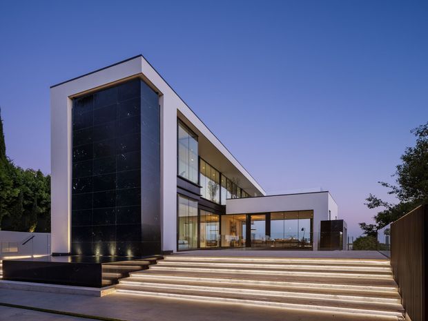 A large contemporary multi-storey residence viewed from the exterior at dusk. The building has a crisp white rendered facade, expansive floor-to-ceiling glazing, a tall dark stone or tile-clad feature wall, and strong horizontal and rectilinear volumes. Broad illuminated steps lead up to the glazed entrance and terrace, with a minimalist landscaped setting and boundary fencing.