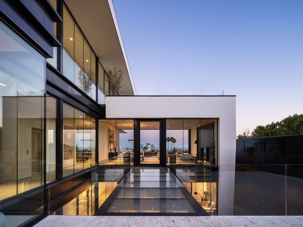 A contemporary multi-level residential building viewed from an elevated exterior terrace or pool deck. The composition features large floor-to-ceiling glazing, dark metal window and door frames, white rendered wall planes, a flat roof, and a reflective dark tiled or polished surface in the foreground. Warm interior lighting is visible through the glazing, with distant urban or coastal views beyond the house.