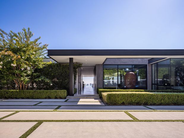 A low-slung contemporary modern house viewed from the front, featuring a broad flat roof with deep overhangs, extensive floor-to-ceiling glazing, dark slender framing, and a covered entry passage. Carefully clipped hedges and geometric concrete-and-grass paving create a formal landscaped approach. A bright interior and reflective glass are visible through the entry and front windows.