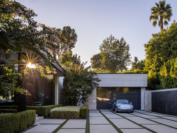 A landscaped residential driveway and contemporary house exterior photographed in warm evening light. Large rectangular concrete pavers separated by narrow planted joints lead to a dark glazed garage door and a white horizontal-clad garage volume. Dense clipped hedges, mature trees, and palms frame the property, while a dark rendered boundary wall runs along the right. A low, flat-roofed home is partially visible behind the planting.