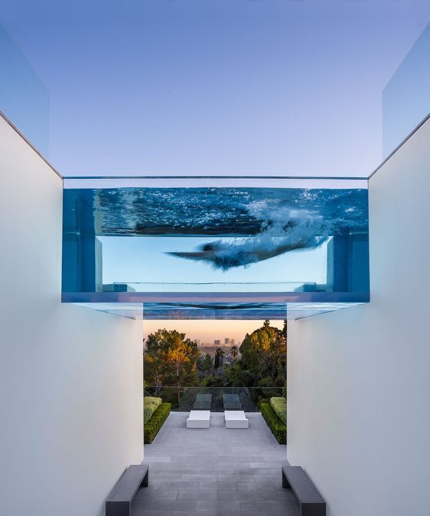 A dramatic modern residential passage with tall white walls and a floating glass-bottom swimming pool spanning overhead. A swimmer is visible through the transparent pool, while broad steps descend toward a landscaped terrace with loungers and a distant urban view. The composition emphasizes clean geometric lines, integrated water architecture, and a strong indoor-outdoor connection.
