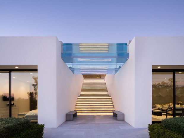 Symmetrical exterior view of a contemporary residence with white rendered walls, large dark-framed glazed openings, and a broad illuminated stairway leading beneath a transparent glass roof or bridge. Interior living spaces are visible through the side glazing, while clipped shrubs and pale paving frame the entrance.