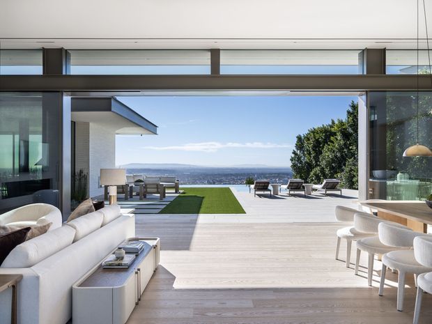A bright, open-plan contemporary living and dining area opening onto a spacious terrace with distant landscape views. The interior features pale timber flooring, floor-to-ceiling glazing, white upholstered seating, a dining table with sculptural chairs, and dark structural framing. The terrace includes outdoor seating and a rectangular lawn area, creating a seamless indoor-outdoor entertaining space.