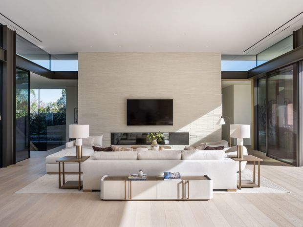 A bright, spacious contemporary living room with a large white sectional sofa arranged around a low sculptural media console and wall-mounted television. The room features pale timber flooring, a full-height light textured feature wall, extensive black-framed glazing, recessed ceiling details, slim built-in shelving, and abundant natural light. A dining or adjacent living area is visible beyond the seating zone.