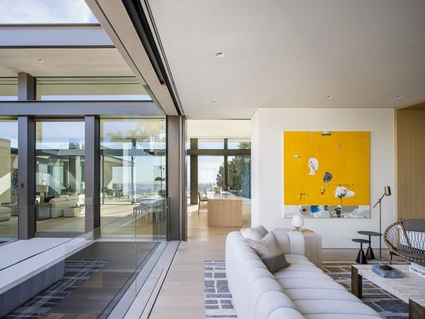 A bright, contemporary living room with a large white sectional sofa, pale timber flooring, patterned area rug, and a vivid yellow abstract artwork. Floor-to-ceiling dark-framed glazing opens toward a spacious terrace or courtyard, creating strong indoor-outdoor connection. The room has a high, clean-lined ceiling and restrained modern furnishings.