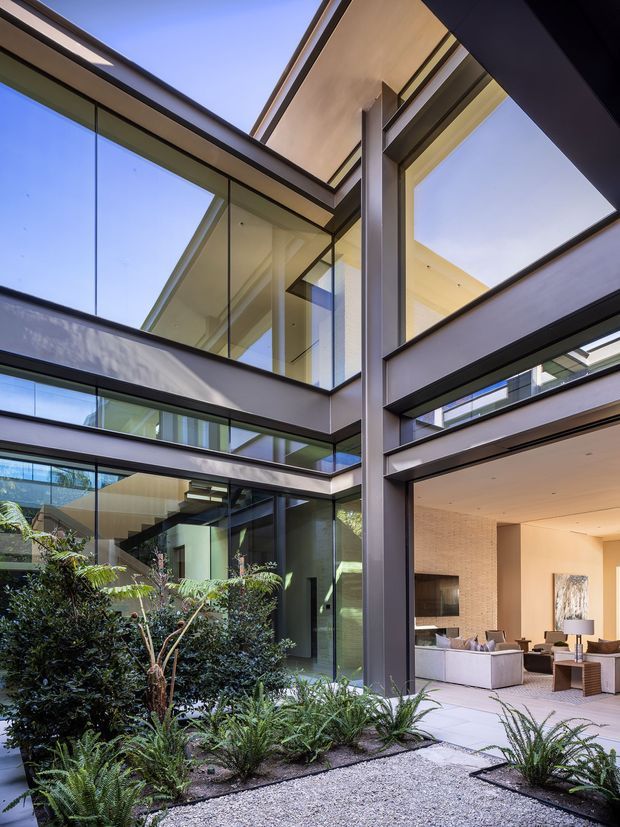 A contemporary multi-level courtyard viewed from an outdoor garden area, with extensive floor-to-ceiling glazing, dark metal structural frames, pale soffits, and open voids that bring daylight into the building. Dense planted beds and pale gravel occupy the foreground, while a refined living room is visible through the glazing on the right. The composition emphasizes indoor-outdoor connection, glazed circulation spaces, and a central landscaped courtyard.