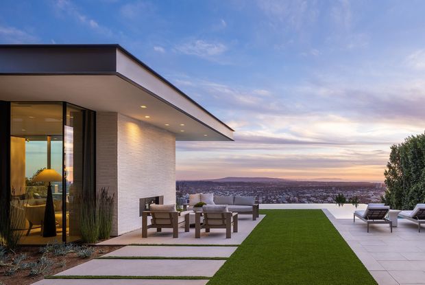 A modern flat-roofed home viewed from its elevated outdoor terrace at dusk. The image shows a minimalist white rendered and textured masonry facade, extensive dark-framed glazing, broad roof overhangs, pale paved outdoor areas, a central strip of lawn, and contemporary outdoor lounge furniture overlooking a distant suburban landscape.