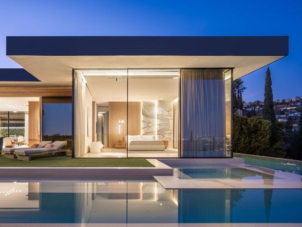Dusk exterior view of a sophisticated modern multi-level residence with strong horizontal roof planes, extensive floor-to-ceiling glazing, warm illuminated interiors, landscaped terraces, and a reflective swimming pool in the foreground. A living area and bedroom are visible through the glazing, with a hillside suburban setting beyond.