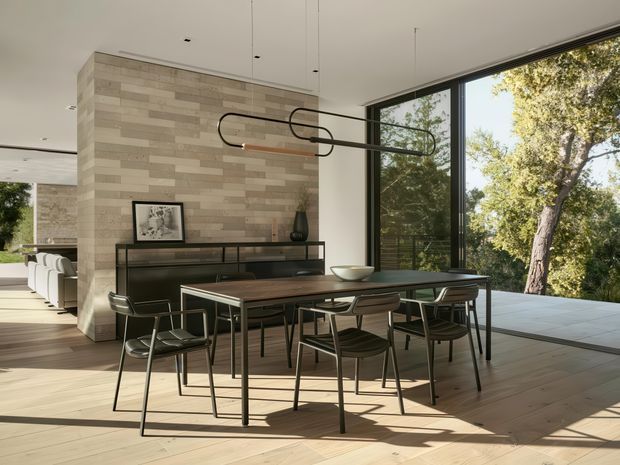 Open-plan contemporary dining area with a long dark timber dining table and black-framed chairs, positioned beside expansive floor-to-ceiling glazing overlooking mature trees. A horizontally clad feature wall, streamlined black sideboard, sculptural linear pendant light, and pale timber flooring create a warm, minimalist modern interior.
